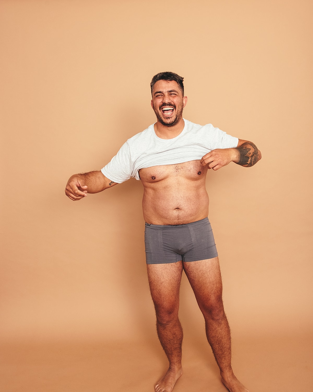 Smiling man lifting shirt against beige background.